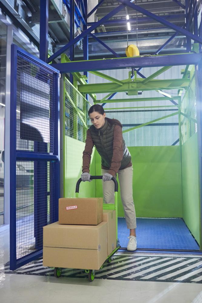Kobieta wjeżdżająca na wózku z paczkami do przemysłowej windy towarowej w magazynie. Otoczenie jest utrzymane w zielono-niebieskiej kolorystyce, a kobieta jest ubrana w roboczy strój.