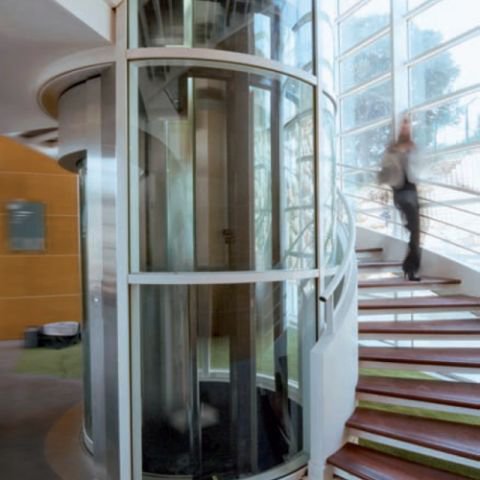 Nowoczesna, przeszklona winda cylindryczna umieszczona obok krętych schodów wewnątrz budynku. W tle widoczne są duże okna, przez które wpada światło, a osoba wchodzi po schodach.