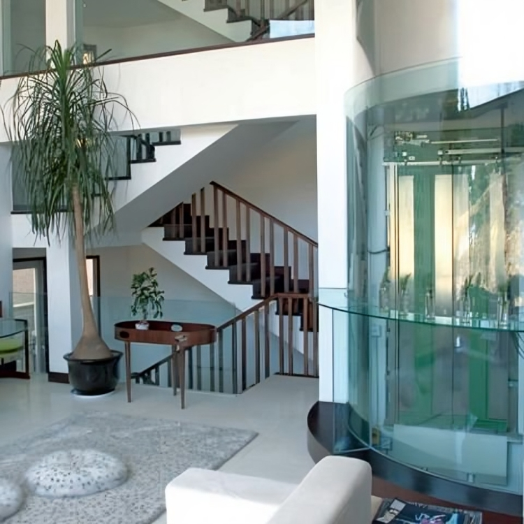 Nowoczesne wnętrze domu z eleganckimi schodami o drewnianych poręczach i przeszkloną, cylindryczną windą po prawej stronie. Przestrzeń jest jasna, z dużymi oknami, roślinami doniczkowymi i minimalistycznym wystrojem.